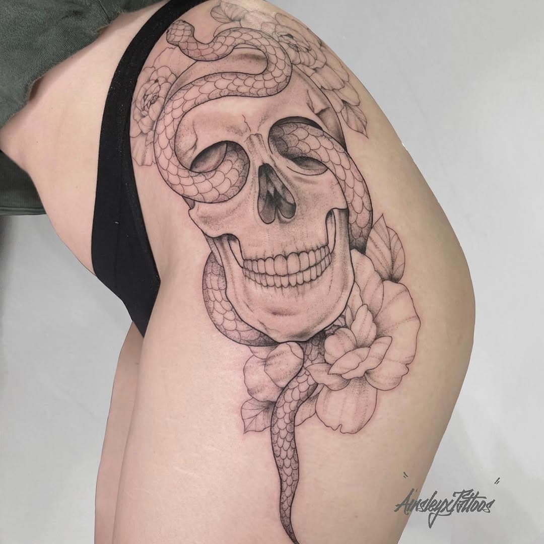 Black-and-grey skull with a serpent threading through the eye sockets and a peony bloom on the thigh by AinsleyXTattoos